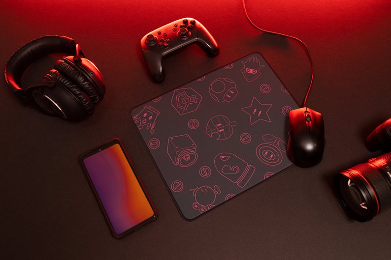 May include: A black gaming mouse pad with a pink outline of various video game characters and symbols. A black wireless controller, a black headset, a black smartphone, and a black gaming mouse are also on the surface.