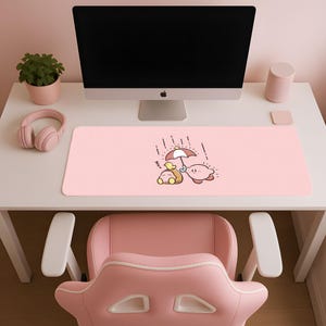 May include: A pink desk mat featuring a cartoon illustration of Kirby and a duck under an umbrella in the rain. The mat is rectangular and sits on a white desk, with a computer monitor, headphones, and a pink chair visible.