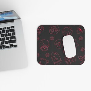 May include: A black mouse pad with a pink outline of video game characters and objects. The mouse pad has a repeating pattern of coins, stars, a mushroom, a shell, a leaf, and a block.