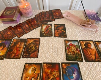 Può includere: Uno stendimento di carte dei tarocchi con una varietà di carte disposte su una tovaglia bianca. Le carte presentano illustrazioni intricate e accenti dorati. Nell'immagine sono visibili anche una candela accesa e incenso.