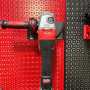May include: A red and gray M18 FUEL brushless grinder with a black handle. The grinder is mounted on a red pegboard. The text "RAPIDSTOP" and "FUEL BRUSHLESS" are visible on the tool.