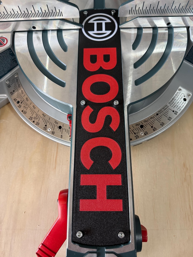 May include: A Bosch mitre saw base featuring a central black plate with the red Bosch logo and a white circular icon. The metallic silver base includes integrated angle scales with black numerical markings and red adjustment levers for precision.