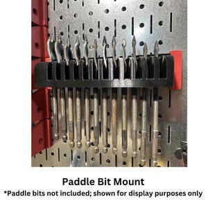 May include: Black paddle bit mount with red accents, holding various metal drill bits. The mount is attached to a metal pegboard. Text on the image reads "Paddle Bit Mount".