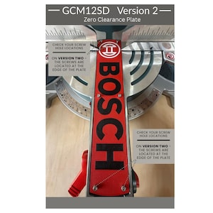 Puede incluir: Una placa de compensación Bosch roja y negra para una sierra GCM12SD Versión 2. La placa tiene el logotipo de Bosch y el texto "COMPRUEBE LAS UBICACIONES DE LOS ORIFICIOS DE LOS TORNILLOS" y "EN LA VERSIÓN DOS - LOS TORNILLOS ESTÁN UBICADOS EN EL BORDE DE LA PLACA".