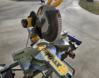May include: A yellow and black DeWalt mitre saw featuring a circular blade guard, a metal base with an integrated angle scale, and a yellow handle. The power tool includes a dust collection port and a metallic fence for material alignment.
