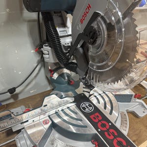 May include: A Bosch miter saw, featuring a silver blade guard and a black and red measuring guide. The saw has a silver base with degree markings and a black handle. The Bosch logo is prominently displayed.