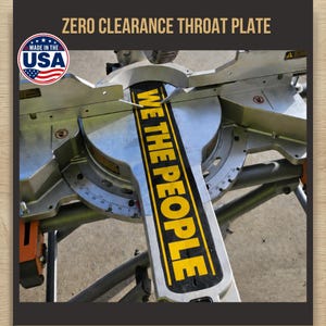 May include: A zero clearance throat plate for a mitre saw, featuring a black surface with the yellow text WE THE PEOPLE printed vertically. The plate is installed on a metallic silver mitre saw base with visible angle markings and a USA flag logo.