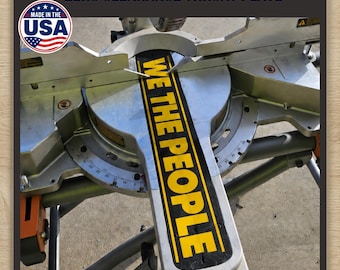 May include: A zero clearance throat plate for a mitre saw, featuring a black surface with the yellow text WE THE PEOPLE printed vertically. The plate is installed on a metallic silver mitre saw base with visible angle markings and a USA flag logo.