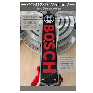 May include: A black Bosch GCM12SD Version 2 zero clearance plate with red lettering and the Bosch logo. The plate features four screw holes located at the edges. Text on the plate reads: GCM12SD Version 2, Zero Clearance Plate, and Check your screw hole locations on version two - the screws are located at the edge of the plate.