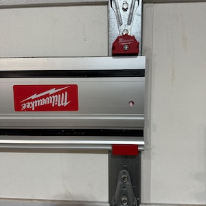 May include: A silver and black Milwaukee tool track system mounted on a white garage door. The track system has a red Milwaukee logo and a red release button.