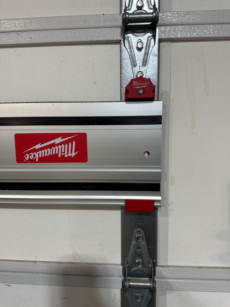 May include: A silver and black Milwaukee tool track system mounted on a white garage door. The track system has a red Milwaukee logo and a red release button.