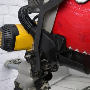 May include: A close-up of a yellow and black DeWalt miter saw. The saw features a large red blade, a black blade guard, and a silver base. The saw is mounted on a white brick wall.