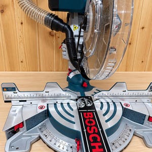 Puede incluir: Una sierra ingletadora Bosch azul y plateada con protector de hoja de plástico transparente, manguera de extracción de polvo negra y base metálica con escalas de medición integradas. La herramienta incluye detalles rojos, etiquetas de advertencia.
