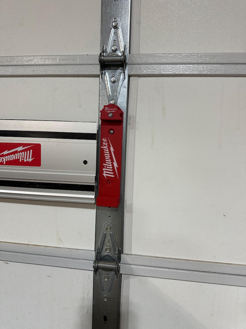 May include: A red Milwaukee magnetic tool holder attached to a garage door track. The holder has a white lightning bolt logo and the word "Milwaukee" printed on it.