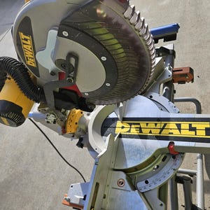 May include: A silver and black DeWalt miter saw with a circular blade. The saw has a yellow and black motor housing and a black dust collection hose. The DeWalt logo is prominently displayed in yellow.