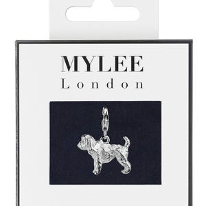 May include: Silver charm of a dog, with a silver clasp, on a black background. The charm is in the shape of a dog, with a textured surface. The charm is packaged in a white box with the words "MYLEE London" printed on it.
