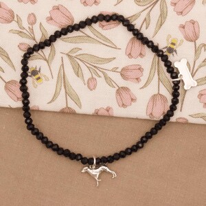 May include: Black beaded bracelet with a silver dog charm and a bone-shaped clasp. The bracelet is displayed on a floral patterned fabric with pink flowers and green leaves.