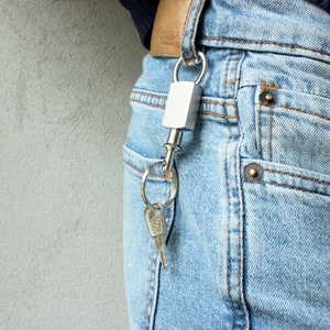 Puede incluir: Un llavero plateado con un candado plateado sujeto a un lazo de cinturón de cuero marrón. El llavero está sujeto a un par de llaves plateadas.