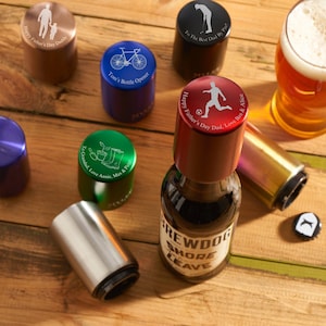 Puede incluir: Surtido de abridores de botellas personalizados en varios colores, incluyendo verde, azul, rojo y dorado. Cada abridor presenta un diseño e inscripción únicos, como una bicicleta, un jugador de fútbol y una escena de jardinería. Los abridores se muestran sobre una superficie de madera con una botella de cerveza y un vaso de cerveza.