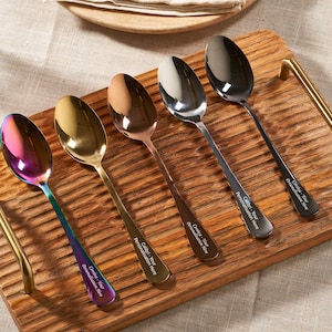 Puede incluir: Cinco cucharas con diferentes acabados metálicos, incluyendo iridiscente, dorado y plateado, se exhiben en una bandeja de madera con asas doradas. Cada cuchara tiene el texto "Calibri - Your Personalization Here" grabado en el mango.