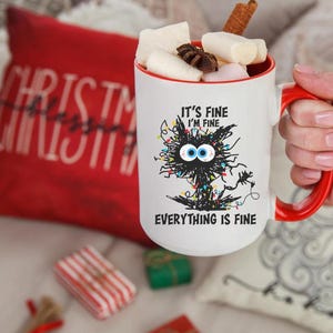 Puede incluir: Taza de cerámica blanca con asa y borde rojos, llena de malvaviscos y una ramita de canela. La taza presenta un gráfico de gato negro con el texto "It's Fine I'm Fine Everything is Fine". Una almohada roja de Navidad está en el fondo.