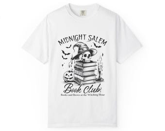 Puede incluir: Camiseta blanca con un gráfico en blanco y negro para el "Midnight Salem Book Club". El diseño incluye una calavera con un sombrero de bruja sobre una pila de libros, una calabaza, murciélagos y una vela encendida. También incluye el texto "Books and Brews at the Witching Hour".
