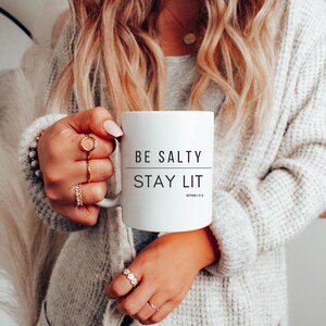 以下が含まれることがあります： 白いセラミック製のマグカップに、黒い文字で「BE SALTY STAY LIT」と書いてあり、その下に小さな文字で「MATTHEW 5:13」と書いてあります。