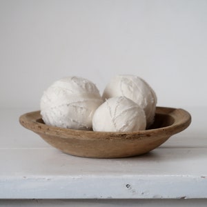 May include: Three white fabric balls in a wooden bowl. The balls are made of white fabric and have a textured surface. The bowl is made of wood and has a natural finish.