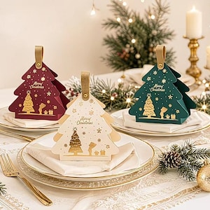Può includere: Tre scatole regalo a forma di albero di Natale in bordeaux, bianco e verde scuro, con dettagli in lamina d'oro, la scritta "Merry Christmas", un grafico di un albero e una piccola maniglia, disposte su piatti con tovaglioli.