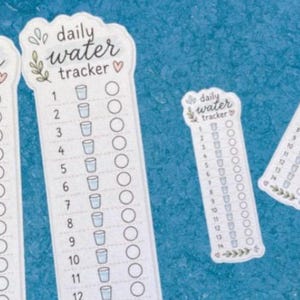 Op de afbeelding: Een verzameling witte rechthoekige stickers met de tekst daily water tracker, genummerde lijsten van 1 tot 14, kleine blauwe bekerpictogrammen en ronde selectievakjes. Elke sticker is versierd met groene bladillustraties en kleine rode hartjes.