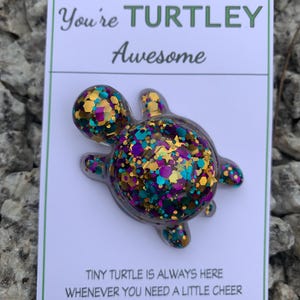 May include: A turtle-shaped pin with gold, teal, and purple glitter, presented on a white card. The card says "You're Turtley Awesome" and "Tiny Turtle is always here whenever you need a little cheer."