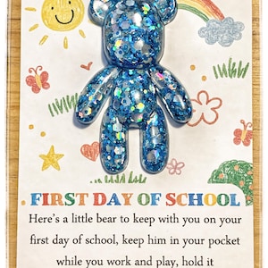 Op de afbeelding: Een blauwe glitterhars berenfiguur bevestigd op een witte kaart met kleurrijke handgetekende illustraties van een zon, regenboog en vlinders. De tekst op de kaart luidt: FIRST DAY OF SCHOOL. Here’s a little bear to keep with you on your first day of school, keep him in your pocket while you work and play, hold it close and remember it’s ok.