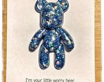 Op de afbeelding: Een blauwe, beervormige harsbedel gevuld met iriserende glitters, gemonteerd op een witte kaart. De kaart bevat de tekst "Worry bear" bovenaan en een gedicht eronder: "I'm your little worry bear, if you feel worried or scared, just hold me tight. I'll keep you safe all day and all night."