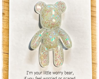 Op de afbeelding: Een in het donker oplichtend berenfiguurtje gevuld met iriserende glitters, bevestigd op een witte presentatiekaart. De kaart bevat de tekst "Worry bear", "GLOW IN THE DARK" en een gedicht over het vasthouden van de beer.