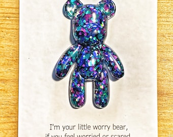 Op de afbeelding: Een kleine beer-vormige figuur gemaakt van heldere hars gevuld met paarse, blauwe en talingkleurige glitters. De beer is bevestigd op een witte kaart met de tekst "Worry bear" en een gedicht: "I'm your little worry bear, if you feel worried or scared, just hold me tight. I'll keep you safe all day and all night."