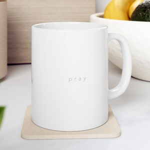 May include: White ceramic mug with the word "pray" printed in a light gray font. The mug sits on a beige coaster. The mug has a curved handle and is in a kitchen setting.
