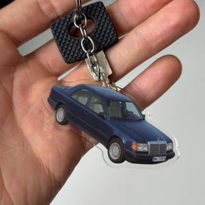 Puede incluir: Un llavero de coche azul con un llavero plateado y un llavero cuadrado negro. El coche es un diseño de sedán clásico con ruedas blancas y una parrilla cromada. El llavero se sostiene en una mano.