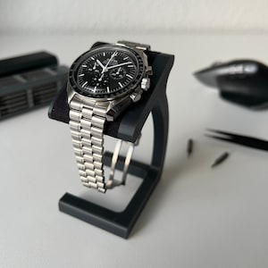 May include: A silver chronograph wristwatch with a black face and a stainless steel band. The watch is on a black watch stand.