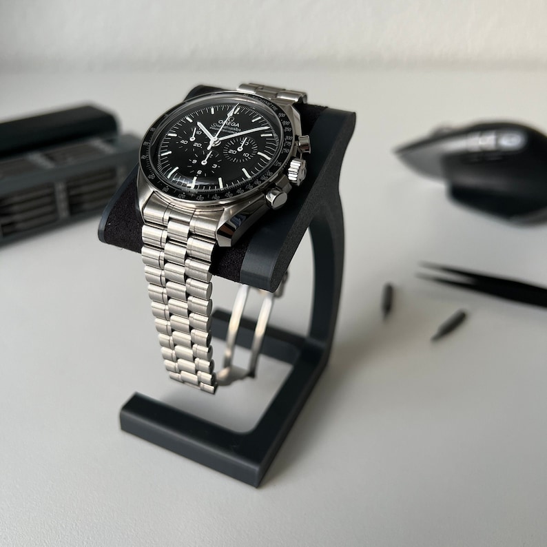 May include: A silver chronograph wristwatch with a black face and a stainless steel band. The watch is on a black watch stand.
