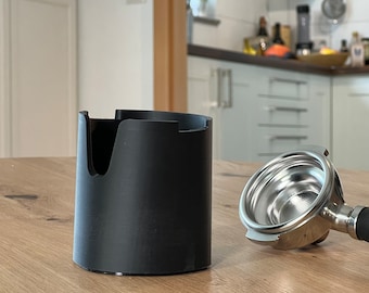 May include: A black plastic coffee grounds container with a silver portafilter on a wooden surface.