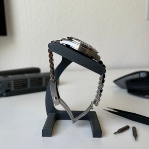 May include: A silver watch with a metal band is displayed on a black watch stand. The watch is on a white surface with three small screwdrivers.