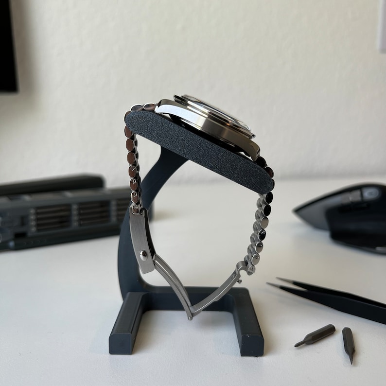 May include: A silver watch with a metal band is displayed on a black watch stand. The watch is on a white surface with three small screwdrivers.