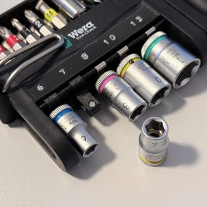Op de afbeelding: Een set Wera-gereedschappen, waaronder doppen en bits, gerangschikt in een zwarte koffer. De doppen zijn zilverkleurig met gekleurde ringen en markeringen. De bits zijn verschillend gekleurd. De afbeelding toont een close-up van de gereedschappen.