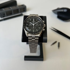 May include: A silver Omega Speedmaster Professional chronograph wristwatch with a black face and a tachymeter bezel. The watch is on a silver metal bracelet and is displayed on a black watch stand.