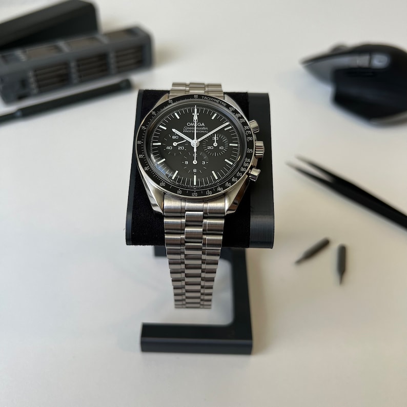 May include: A silver Omega Speedmaster Professional chronograph wristwatch with a black face and a tachymeter bezel. The watch is on a silver metal bracelet and is displayed on a black watch stand.