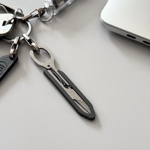 Op de afbeelding: Een compact, opvouwbaar metalen schaartje bevestigd aan een zilveren sleutelhanger. De schaar heeft een matzwarte handgreep en een metalen mesmechanisme, ontworpen om in een slanke, langwerpige vorm te vouwen voor gemakkelijke draagbaarheid.