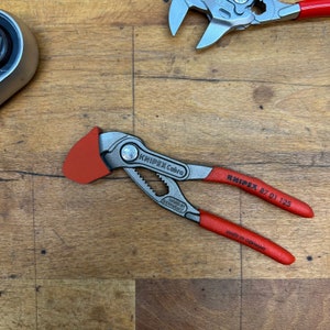 Puede incluir: Un alicate Knipex Cobra con mangos rojos y una tapa protectora roja. Los alicates son plateados y tienen el texto "KNIPEX Cobra" y "MADE IN GERMANY" impresos. Otra llave inglesa es visible en el fondo.