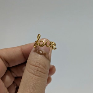 May include: A gold-coloured wire ring spelling out the word "love" in a cursive font. The ring is held between two fingers against a plain, light background. The ring is a delicate piece of jewellery.