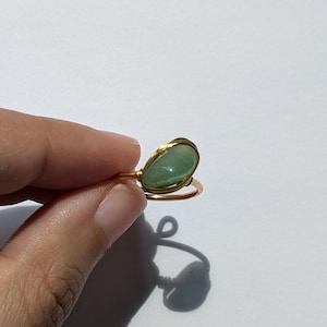 May include: A gold-coloured wire ring with a teardrop-shaped, light green gemstone. The gemstone is set in a gold-coloured bezel. The ring is held against a white background, highlighting the ring's design and colour.