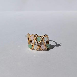 May include: A gold-tone wire ring featuring a geometric pattern. The ring is embellished with small, round beads in pink, green, and clear hues. The ring sits on a white surface, with a shadow to the right.
